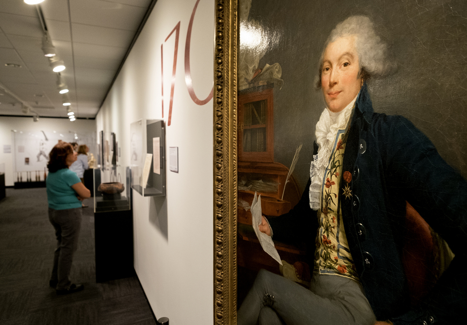 A closeup of a 1791 oil painting of Le Marquis D’Acqueville by Aimée Duvivier, while in the background a visitor examines a document in the "Deep in the Vaults of Texas" exhibition at the LBJ Library.